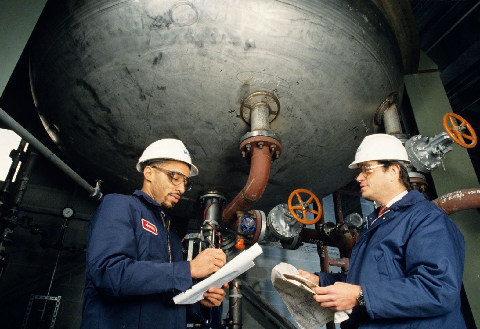 Two engineers in hard hats inspecting piping beneath a large industrial vessel.